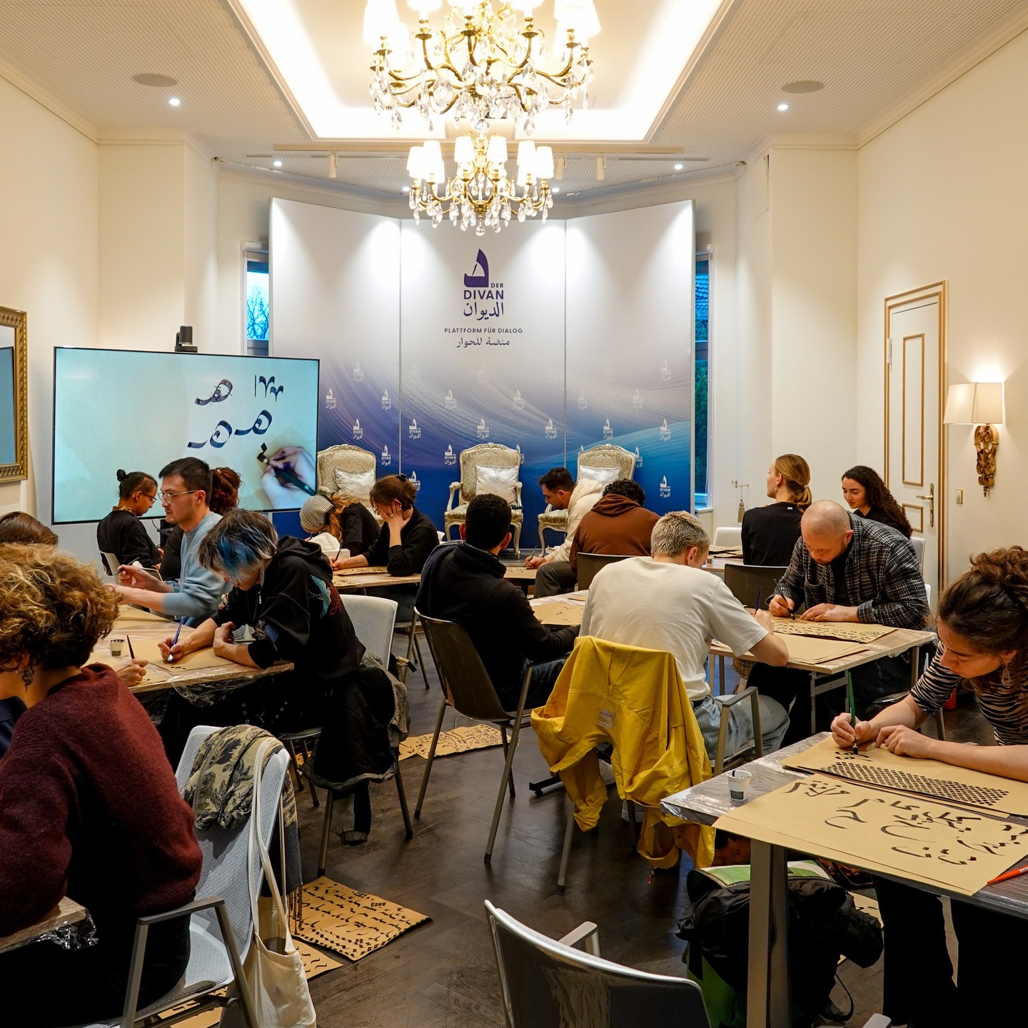 Am 9. Dezember 2025 fand im Divan – Das Arabische Kulturhaus ein Kalligraphie-Workshop mit Daniel Arab in Kooperation mit der Humboldt-Universität zu Berlin statt. Die Veranstaltung bot Raum für eine praktische Auseinandersetzung mit arabischer Schrift als kultureller Ausdrucksform und als Medium des Austauschs. In zwei aufeinanderfolgenden Kursen diskutierten und erprobten die Teilnehmenden Schriftstile, historische Bezüge und ästhetische Prinzipien im Kontext eines interkulturellen Dialogs.
أُقيمت في 9 كانون الأول/ديسمبر 2025 في الديوان – البيت الثقافي العربي ورشة للخط العربي بإشراف دانيال عرب وبالتعاون مع جامعة هومبولت في برلين. شكّلت الورشة مساحة للتعامل العملي مع الخط العربي بوصفه تعبيرًا ثقافيًا ووسيلة للحوار. وعلى مدار دورتين متتاليتين، ناقش المشاركون وجربوا أنماط الخط، وسياقاته التاريخية، وجمالياته ضمن إطار حوار ثقافي.
#ArabischeKalligraphie #ArabicCalligraphy #DivanBerlin #HUBerlin #KunstUndKultur #DeutschArabisch Am 9. Dezember 2025 fand im Divan – Das Arabische Kulturhaus ein Kalligraphie-Workshop mit Daniel Arab in Kooperation mit der Humboldt-Universität zu Berlin statt. Die Veranstaltung bot Raum für eine praktische Auseinandersetzung mit arabischer Schrift als kultureller Ausdrucksform und als Medium des Austauschs. In zwei aufeinanderfolgenden Kursen diskutierten und erprobten die Teilnehmenden Schriftstile, historische Bezüge und ästhetische Prinzipien im Kontext eines interkulturellen Dialogs.
أُقيمت في 9 كانون الأول/ديسمبر 2025 في الديوان – البيت الثقافي العربي ورشة للخط العربي بإشراف دانيال عرب وبالتعاون مع جامعة هومبولت في برلين. شكّلت الورشة مساحة للتعامل العملي مع الخط العربي بوصفه تعبيرًا ثقافيًا ووسيلة للحوار. وعلى مدار دورتين متتاليتين، ناقش المشاركون وجربوا أنماط الخط، وسياقاته التاريخية، وجمالياته ضمن إطار حوار ثقافي.
#ArabischeKalligraphie #ArabicCalligraphy #DivanBerlin #HUBerlin #KunstUndKultur #DeutschArabisch